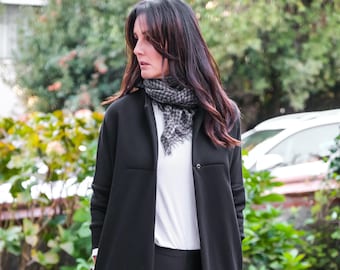 May include: A woman in a black coat, black leggings, a white top, a grey and white scarf, and black boots. The coat features a button and pockets. The background shows greenery and a car.