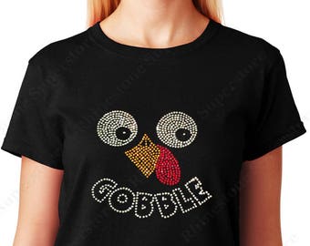 May include: Black t-shirt with a rhinestone turkey face design. The turkey face features white eyes, a yellow and orange beak, a red tongue, and the word "GOBBLE" in white rhinestones. A fun, festive top for Thanksgiving.