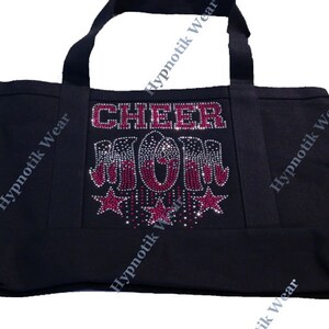 May include: Black canvas tote bag with the words "CHEER MOM" in pink rhinestones. The letters are decorated with silver and red rhinestones. The bag has two black handles.