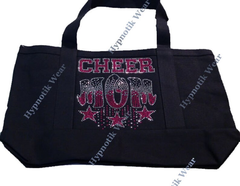 May include: Black canvas tote bag with the words "CHEER MOM" in pink rhinestones. The letters are decorated with silver and red rhinestones. The bag has two black handles.