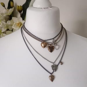 May include: A selection of layered necklaces with heart-shaped pendants in gold, silver, and bronze. The necklaces are displayed on a white mannequin bust, highlighting the different chain styles and pendant designs. The background is a soft, neutral colour.