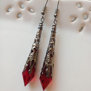 May include: A pair of dangle earrings with a dark metal filigree design. Each earring features a faceted, deep red crystal pendant. The earrings are suspended from a simple hook.