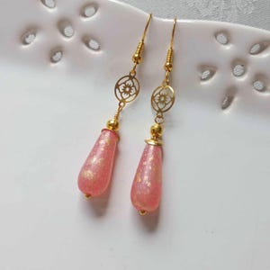 May include: A pair of gold dangle earrings with pink teardrop-shaped beads. The beads have a subtle shimmer and are accented with gold flecks.