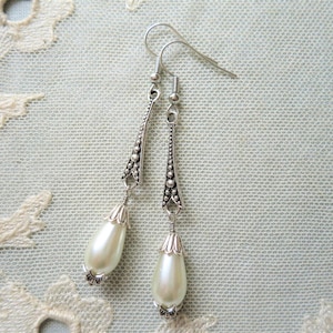 A pair of handmade art deco style dark silver and off white coloured metal and glass pearl drop bead long tapered earrings with raised dot detail lying on a cream lace doily