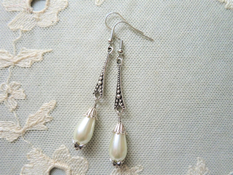 A pair of handmade art deco style dark silver and off white coloured metal and glass pearl drop bead long tapered earrings with raised dot detail lying on a cream lace doily