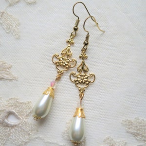 A pair of handmade baroque style gold and off white coloured long drop earrings with ornate openwork floral detail, tiny crystals and pearly teardrop dangle lying on a cream lace doily