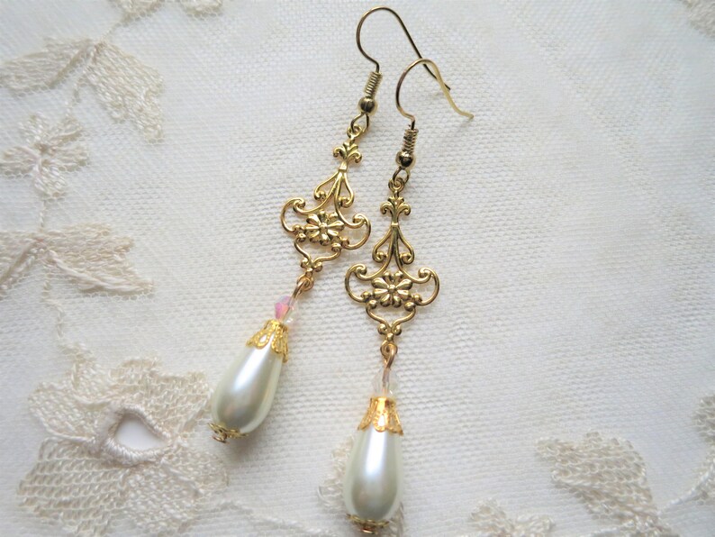 A pair of handmade baroque style gold and off white coloured long drop earrings with ornate openwork floral detail, tiny crystals and pearly teardrop dangle lying on a cream lace doily