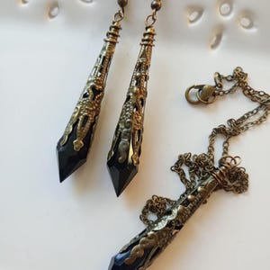 May include: A jewelry set featuring a necklace and matching earrings. The earrings and pendant have a bronze-colored filigree design with a black, pointed crystal. The necklace has a bronze chain and clasp.