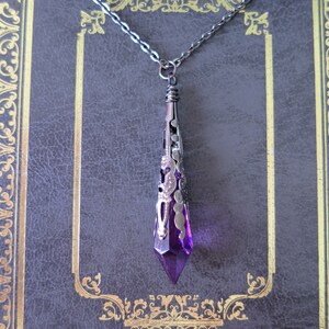 A handmade purple and gunmetal coloured ornately stamped metal and acrylic faceted pointed teardrop pendant necklace on chain lying on a grey faux leather diary embossed with an intricate gold border