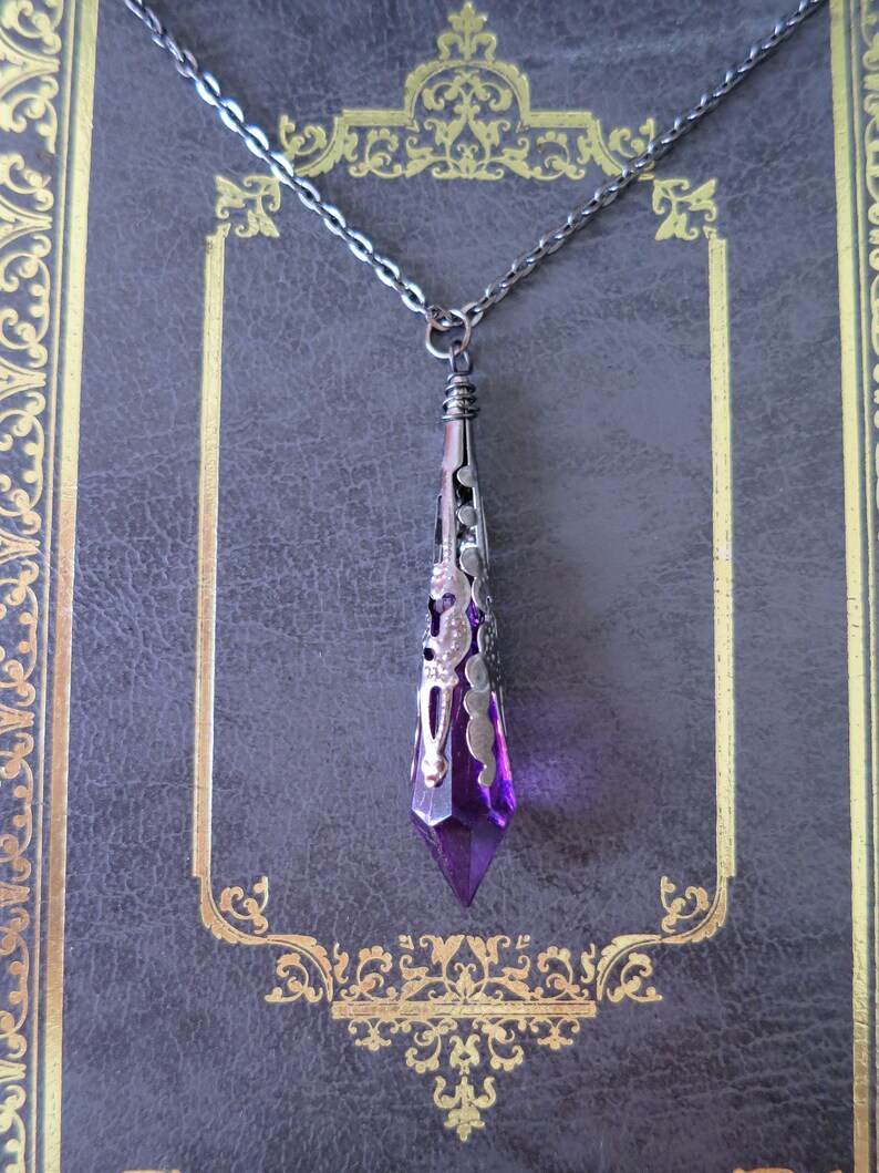 A handmade purple and gunmetal coloured ornately stamped metal and acrylic faceted pointed teardrop pendant necklace on chain lying on a grey faux leather diary embossed with an intricate gold border