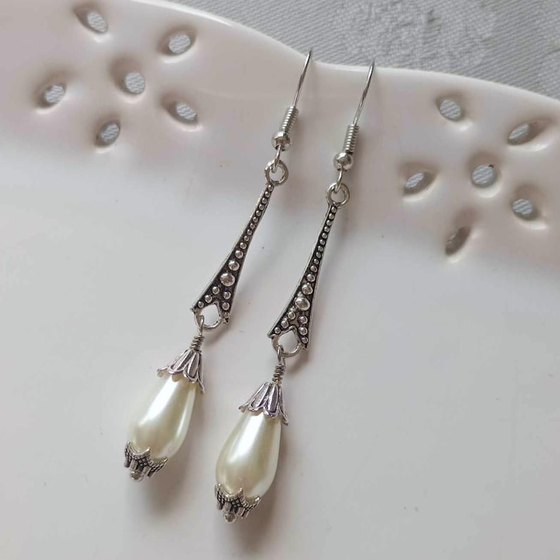 Pair of handmade 1920s Style Elegant Teardrop earrings featuring antiqued silver tapered bars with raised dot detail and white ivory teardrop glass pearls, silver fluted bead caps & wires displayed hooked onto the rim of a white decorative ceramic dish.