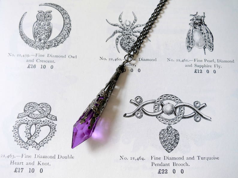 A handmade transparent purple and gunmetal coloured ornate metal and acrylic drop bead pendant necklace on chain lying on an antique jewellery catalogue page