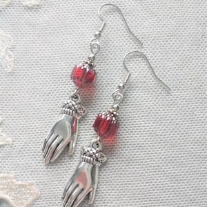 A pair of handmade antique style dark silver and transparent ruby red coloured metal and faceted glass bead earrings with human gloved hand shaped charm dangle laying on a cream lace doily