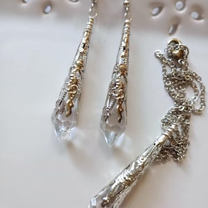 以下が含まれることがあります： ネックレスとイヤリングのジュエリーセット。イヤリングとペンダントは、下部に透明なファセットクリスタル、シルバーカラーのメタルフィリグリー、ゴールドカラーのアクセントが付いています。ネックレスはシルバーチェーンです。