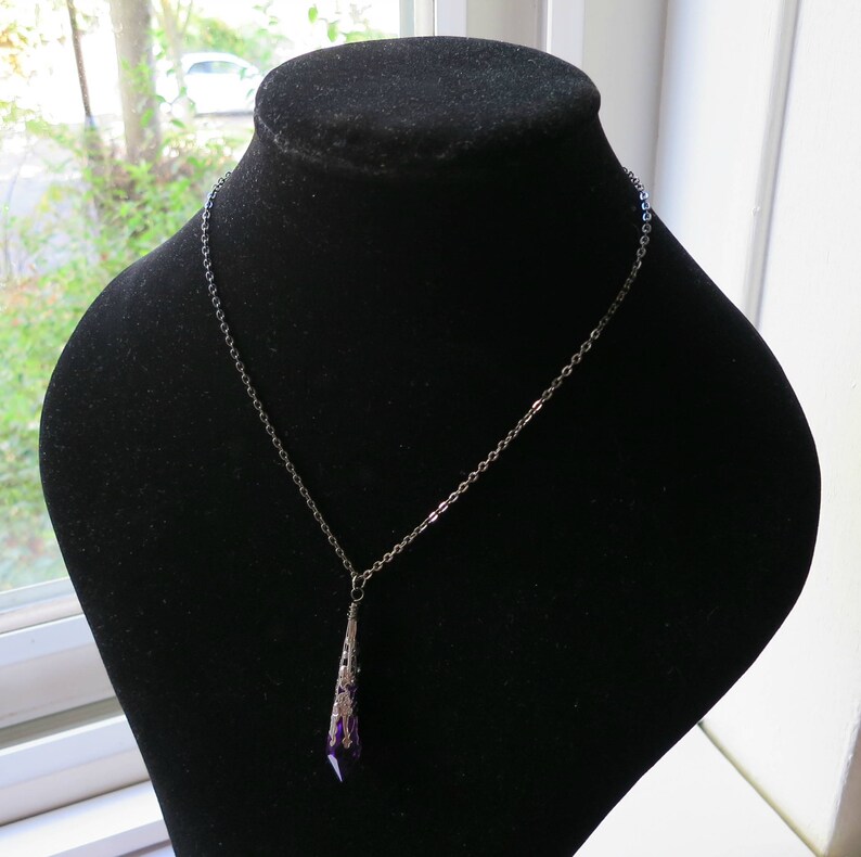 Pu&ograve; includere: Una collana con catena d'argento con un ciondolo di cristallo viola. Il ciondolo ha la forma di una lacrima ed &egrave; decorato con un motivo a filigrana d'argento.