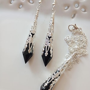 May include: A silver-toned jewelry set featuring a necklace and matching earrings. The set includes a pendant and earrings with black, pointed crystal-like stones encased in ornate, filigree metalwork. The earrings hang from silver hooks.