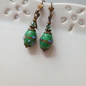 May include: A pair of green dangle earrings with an oval shape. The earrings have a bronze-colored metal with green and blue accents. The earrings are on a white surface.