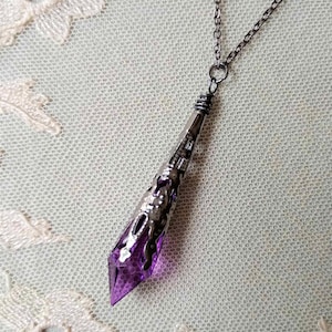 A handmade transparent purple and gunmetal coloured ornate metal and acrylic long pointed drop bead pendant necklace laying on a cream lace doily