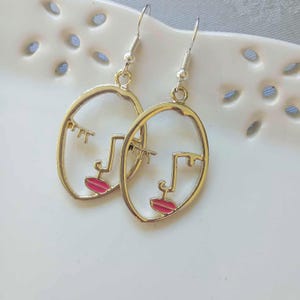 以下が含まれることがあります： 顔のピンク色の輪郭が描かれた、ゴールドトーンのイヤリングのペア。イヤリングは円形で、内側に顔が描かれています。