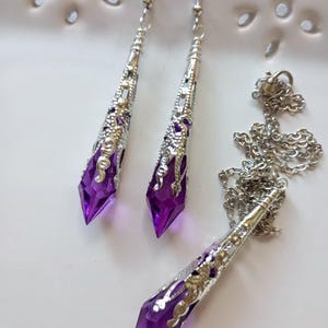 以下が含まれることがあります： ネックレスとイヤリングのジュエリーセット。イヤリングとペンダントは、紫色のクリスタルのような中心部を持つ銀色のフィリグリーデザインです。セットは白い表面に表示されています。