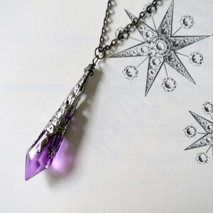 Pu&ograve; includere: Una collana con un ciondolo d'argento e una punta di cristallo viola. Il cristallo &egrave; sfaccettato e ha una montatura in argento. La collana &egrave; su uno sfondo bianco.