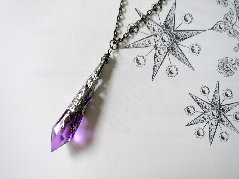 Pu&ograve; includere: Una collana con un ciondolo d'argento e una punta di cristallo viola. Il cristallo &egrave; sfaccettato e ha una montatura in argento. La collana &egrave; su uno sfondo bianco.