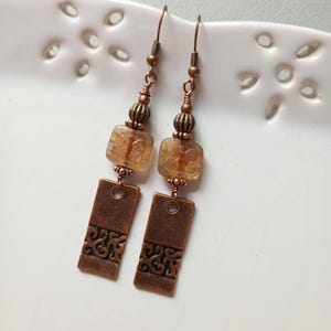 May include: A pair of handcrafted copper-toned earrings. Each earring features a rectangular pendant with a decorative cutout design, a square amber-colored bead, and smaller round beads. The earrings hang from copper-colored ear wires.