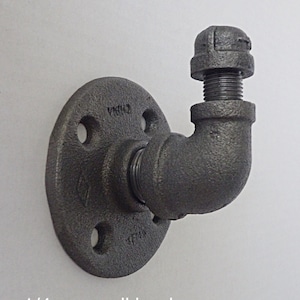 Puede incluir: Un gancho de pared de metal negro con una conexión de tubería de 1/4 de pulgada y una tapa. El gancho está unido a una placa redonda de metal con tres agujeros.