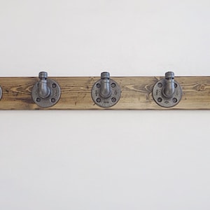 Könnte beinhalten: Ein rustikaler Holz-Kleiderständer mit fünf Metallhaken. Die Haken sind aus Rohrverbindungen gefertigt und haben eine silberne Oberfläche. Das Holz ist dunkelbraun.