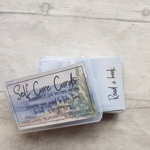 May include: A set of self-care cards in a clear plastic box with a watercolour design. The box has the text "Self Care Cards" and "Prompts to give you some me time because you need to take time to look after yourself". The box also says "Contains 36 cards - one for every day of the month & more".
