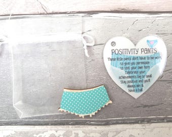 May include: A small teal wooden cutout shaped like underwear with white polka dots, accompanied by a heart-shaped tag reading POSITIVITY PANTS and a poem about celebrating achievements, all presented with a sheer white drawstring gift bag.