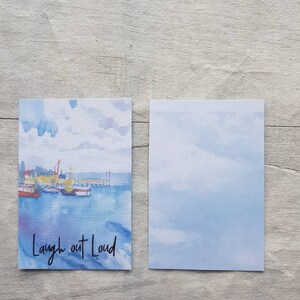 May include: A watercolour painting of a harbour scene with boats and a pier. The painting is on a white card with the text "Laugh out Loud" written in black ink.