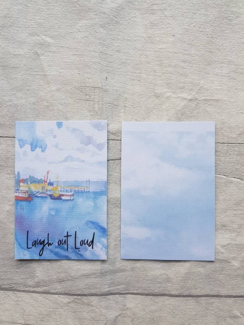 May include: A watercolour painting of a harbour scene with boats and a pier. The painting is on a white card with the text "Laugh out Loud" written in black ink.
