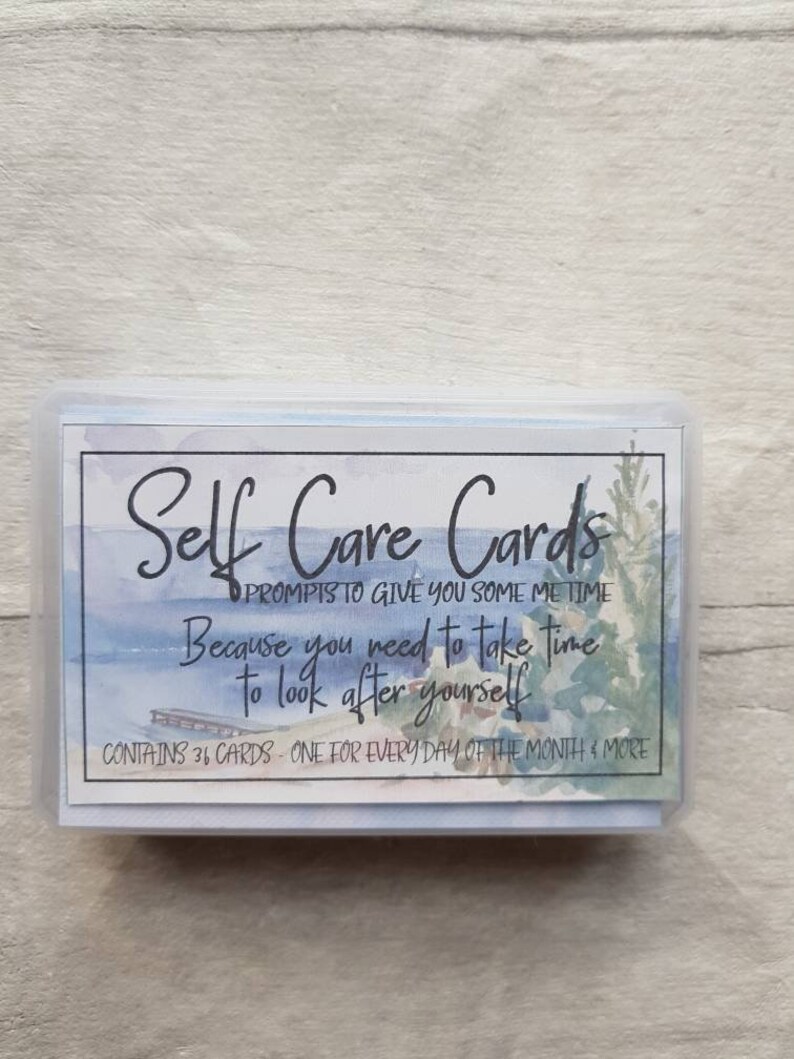 May include: A box of self-care cards with a watercolour design of a lake and a jetty. The text on the box reads "Self Care Cards" and "Prompts to give you some me time. Because you need to take time to look after yourself." The box also states that it contains 36 cards, one for every day of the month and more.