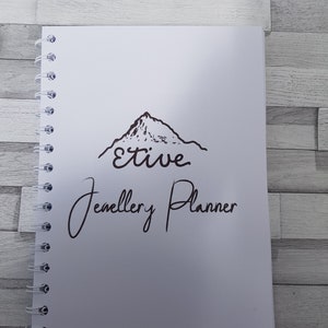 May include: A white spiral bound notebook with a black mountain illustration and the text "Etive Jewellery Planner".