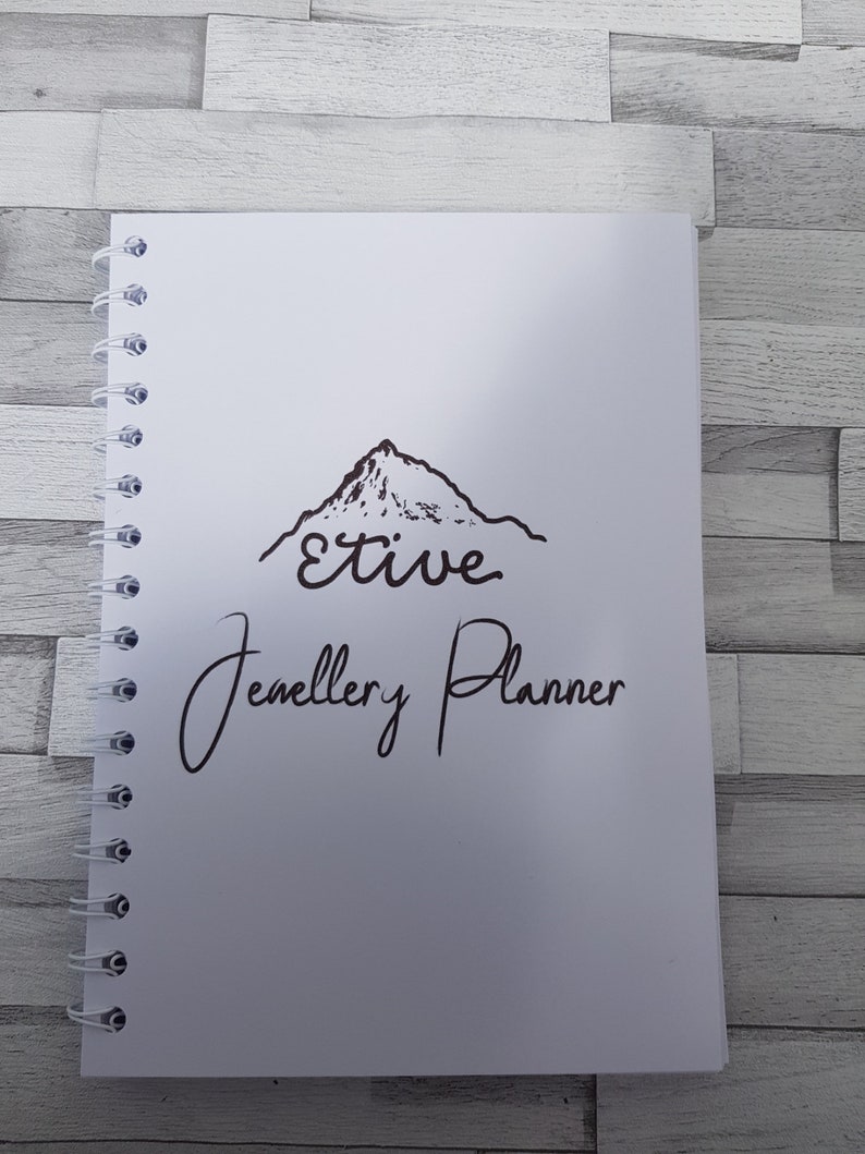 May include: A white spiral bound notebook with a black mountain illustration and the text "Etive Jewellery Planner".