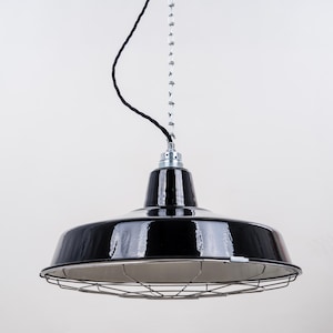Op de afbeelding: Zwarte hanglamp met een glanzende afwerking en een draadkooi. De lamp hangt aan een ketting en een zwarte kabel tegen een witte achtergrond. Een moderne industriële lamp.