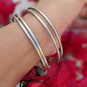 May include: Three silver bangles are worn on a wrist, reflecting light. The bangles are smooth and round, with a polished finish. The background is blurred, featuring red flowers.
