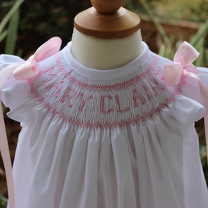 May include: White baby dress with pink embroidered lettering that says "Mary Claire" and pink satin ribbon ties.