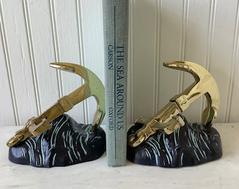 May include: A pair of gold anchor bookends with black and green bases. The bookends are holding up a book titled "The Sea Around Us" by Rachel Carson.