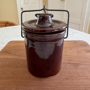 May include: A dark brown ceramic crock with a lid and a metal handle. The cylindrical container has a glossy finish and sits on a wooden surface. The lid has a small knob for easy removal. A classic kitchenware item.