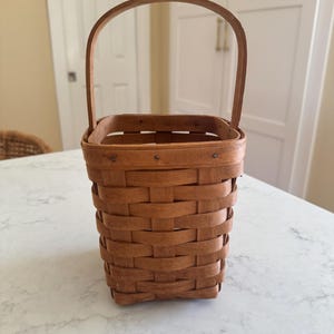 May include: A small, rectangular brown woven basket with a curved handle. The basket is made of thin strips of wood woven together in a basket weave pattern. The handle is attached to the top of the basket with small metal rivets. The basket is empty.