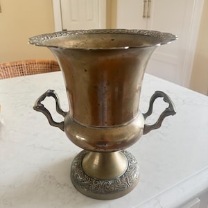 May include: A metallic urn-shaped vessel with a wide, scalloped rim and two ornate, curved handles on the sides. The body features a smooth, tarnished brass finish, while the base and rim are decorated with intricate, embossed silver-toned patterns.