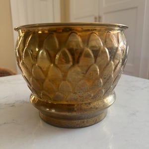 May include: A round brass planter featuring a textured exterior with a repeating embossed scale pattern. The metal surface displays a warm, metallic gold tone with subtle signs of natural aging and patina throughout the rounded body.