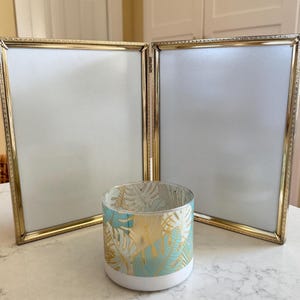 May include: A gold-toned tri-fold picture frame with a white background and a decorative glass candle holder with a teal and gold leaf pattern.