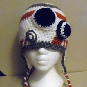 Może przedstawiać: Biały, czerwony i szary kapelusz na szydełku z czarnymi i białymi detalami oczu. Kapelusz ma długie sznurki, które są białe, czerwone i szare.