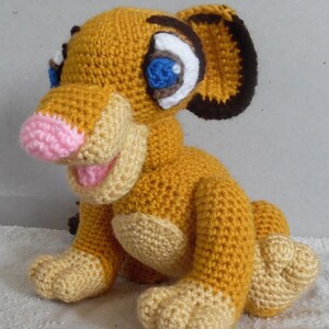 May include: A crocheted stuffed lion cub toy with a pink nose, blue eyes, and a yellow and tan color scheme.