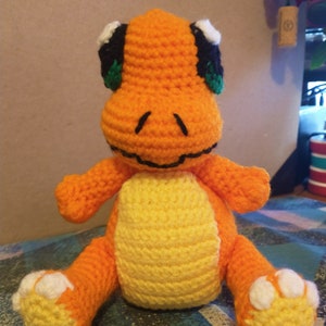 May include: A crocheted orange and yellow dinosaur toy with green eyes and white accents.