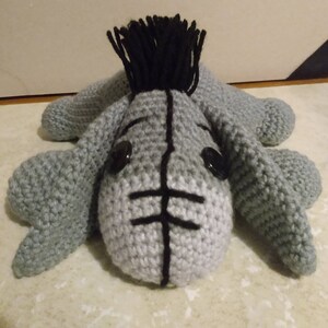 May include: A gray crocheted stuffed animal resembling a donkey with black yarn for the mane and eyes. The donkey has a black stitched mouth and nose.