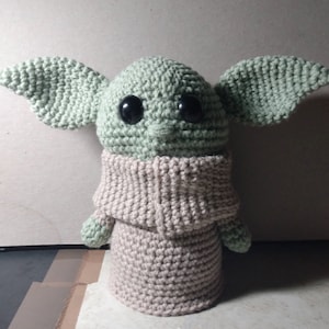 May include: A crocheted plush toy of a green character with large black eyes and a beige knitted sweater. The character has large ears and a small mouth.